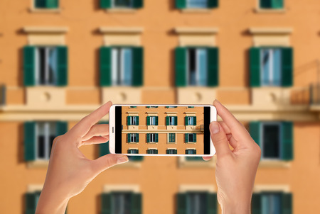 A tourist is taking a photo of part of the facade of an orange italian building with windows with green shutters on a mobile phoneの写真素材