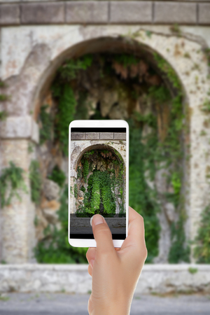 A man is making a photo of a small cave overgrown with ivy near Terrazza del Pincio. Rome, Italy on a mobile phoneの写真素材