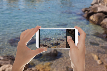 A tourist is taking a photo of a small part of shingle beach with clear water on the Amalfi coast on a mobile phoneの写真素材