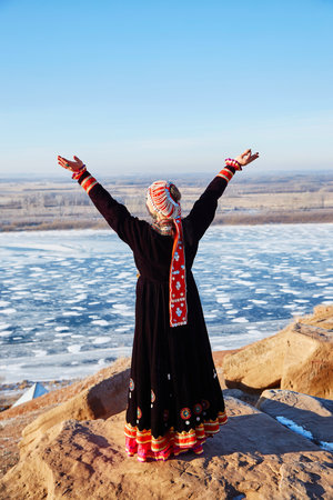 Back view of anonymous female in traditional dress and hat raising arms and enjoying sunlight with closed eyes while standing on cliff against frozen river and cloudless blue skyの写真素材
