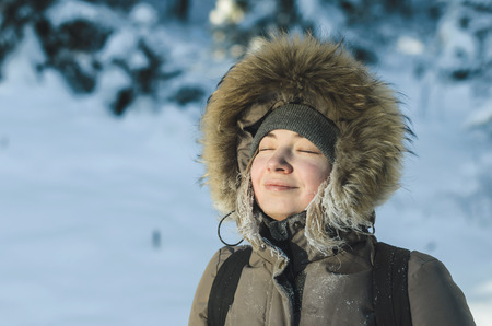 Girl in warm winter jacket with a hood with fur, eyes closed, smiling, enjoying the beautiful weather.の写真素材