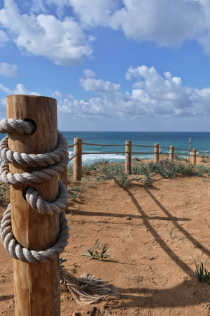 Rope fence on the beach with blue sky and sea in background in Netanya. Sturdy rope fence, composed of thick, gray ropes secured between light brown wooden postsの写真素材