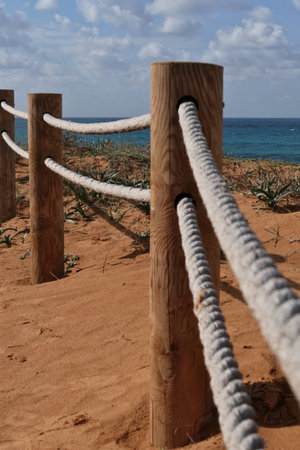 Rope fence on the beach with blue sky and sea in background in Netanya. Sturdy rope fence, composed of thick, gray ropes secured between light brown wooden postsの写真素材