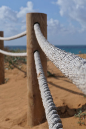 Rope fence on the beach with blue sky and sea in background in Netanya. Sturdy rope fence, composed of thick, gray ropes secured between light brown wooden postsの写真素材