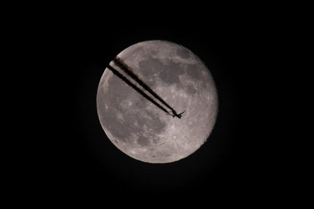 The moon and an airplane in the night sky. The plane is flying across the moon, against a black...
