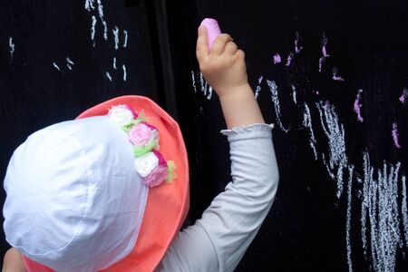 little girl child in a hat draws with chalk on a black fenceの写真素材