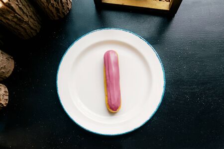 Eclair with pink glaze on a white plate. Raspberry eclair in the glaze on a black wooden table. Top view.の写真素材