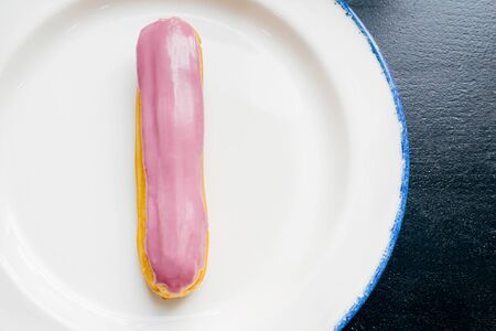 Eclair with pink glaze on a white plate. Raspberry eclair close up in the glaze on a black wooden table. Top view.の写真素材