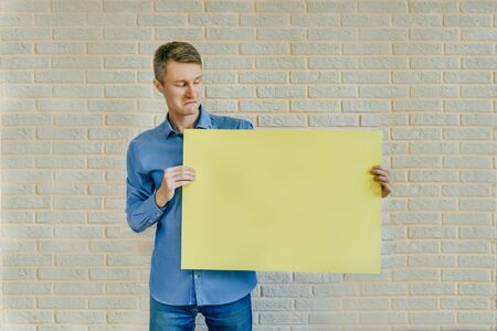 Young man look into empty yellow banner and light brick background. Surprised man holding...