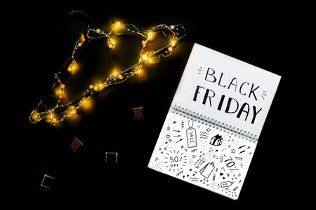 Black friday flat lay with note and black shoulder with garland on black background. Lettering Black friday and Doodle in note.の写真素材