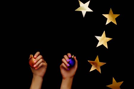 Beautiful christmas composition with gold stars. Gold christmas ball in hand with coral manicure and violet ball in hand with violet manicure on black background. Flat lay design. Copy Space.の写真素材