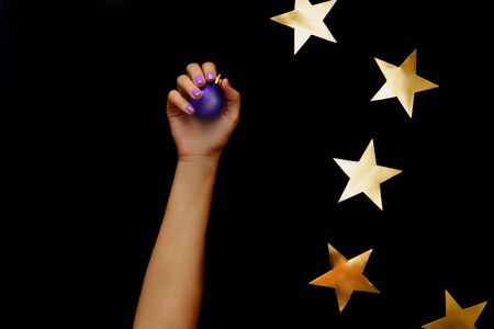 Beautiful christmas composition with gold stars. Violet christmas ball in hand with violet manicure on black background. Flat lay design.の写真素材