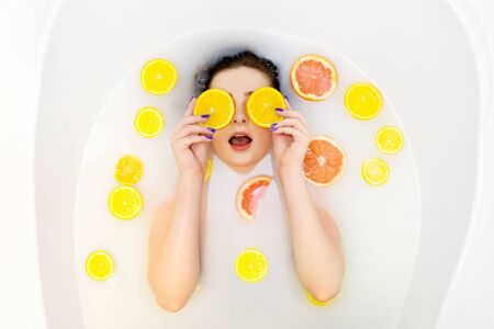 Surprised girl in bath with milk and orange, lemon, grapefruit slices. Top view, orange slices in...