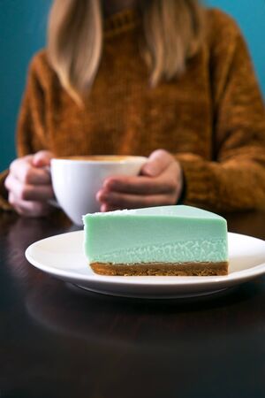 Woman in terracotta sweater headless and hands with latte. Turquoise background, brown wooden table. Foreground mint cheesecake.の写真素材