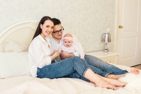 Happy young father, mother and cute baby girl lying on light bed.