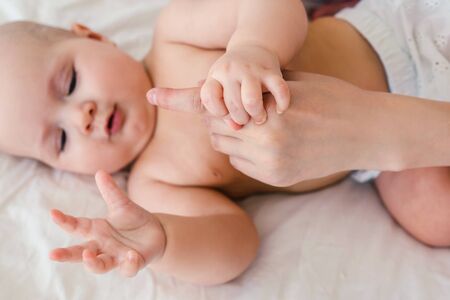 Naked cute baby girl lie on a light background in bed and hold moms finger. At home.