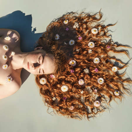 Close up portrait of young ginger curly girl with flowers in hair above. Top view. Concept of spring...