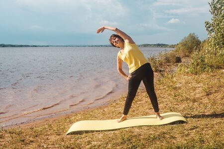 Senior woman making yoga and fitness near river in summer. Lockdown concept, sport alone.