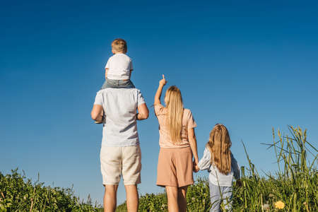 Happy family of mother, father and two children have a rest in field. Mother shows a finger to the sky and they looks up. Back view.の写真素材