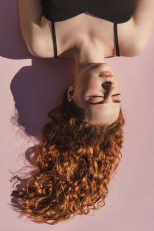 Curly ginger girl with closed eyes upside down on pink background. Tenderness, beauty, spa, hair care concept.の写真素材