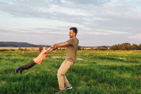Father circles son around him, playing together. Blue sky background, green field, front view.
