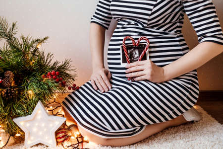 Close up of cropped pregnant woman making a heart from lollipops and ultrasound inside on her belly. Christmas decor on background.の写真素材
