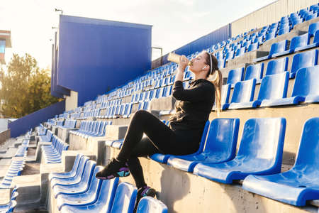 Fitness woman in black tracksuit sitting on the podium of the stadium and drink water. Woman after training. Sunset light, full length.の写真素材