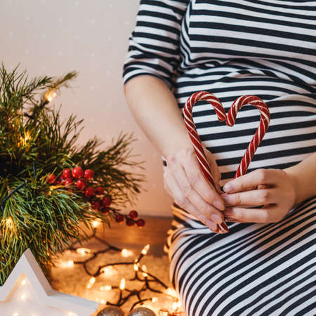 Close up of cropped pregnant woman making a heart from lollipops on her belly. Christmas decor on background.の写真素材
