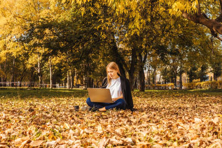 Young businesswoman working with laptop on nature with coffee. Autumn park, sunny day.の写真素材