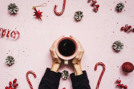 Female hands holding cup of coffee on christmas pink sparkling background. Festive concept for holidays. Flat lay style, greeting card. Red baubles and pine cones as frame around hands.の写真素材