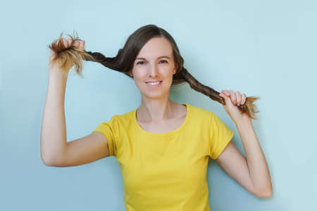 Joyful excited smiling young woman with long hair in hands on blue background. Yellow t-shirt. Funny...