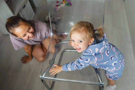 Funny little girl in beautiful suit at home on transparent chair. Mother look at daughter and smile. Light interior.の写真素材