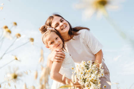 Beautiful young mother and her daughter having fun at the field. Chamomile on foreground, sky background. White clothes.の写真素材