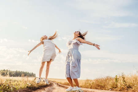Beautiful young mother and her daughter having fun and jumping at the country road. Ground on foreground, sky background. White clothes, freedom concept.の写真素材