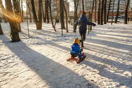 A man run and rolls a small child boy on a snowmobile through the snow in the park. Back view.の写真素材