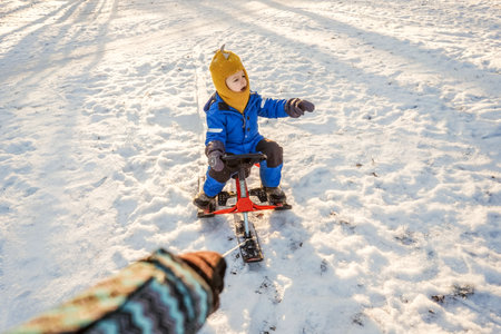 Womans hand rolls a small child boy on a snowmobile through the snow in the park. Sun light and trees shadows on background.の写真素材