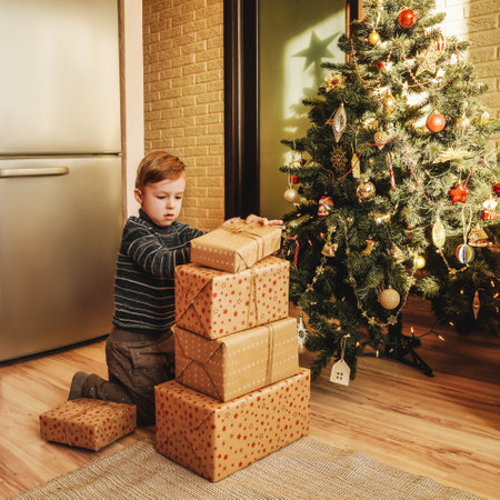 A little boy builds big stack of Christmas presents. Christmas tree on the background. New Years and Christmas concept.の写真素材