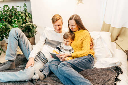 Happy family mother, father and boy at home in bed watch video in tablet. Green houseplants around, sustainable design. Yellow and gray colors 2021 year.の写真素材