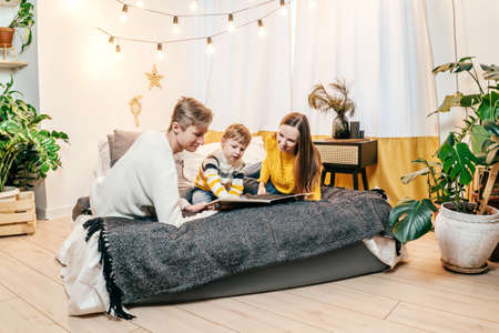 Happy family mother, father and boy at home in bed read the book. Green houseplants around, sustainable design. Yellow and gray colors 2021 year.の写真素材
