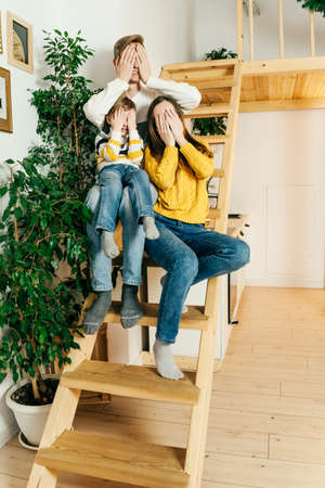 Happy family mother, father and boy at home sit on stairs and close eyes with hands. Green houseplants around, sustainable design. Yellow and gray colors 2021 year.の写真素材