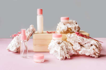 Blank white bottles with cream and lotion on wooden brick, white stones around. Natural organic cosmetics, sustainable lifestyle concept. Front view, copy space. Pink blue background.の写真素材