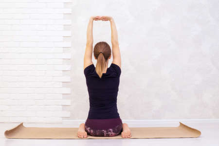 Portrait of beautiful young woman practicing yoga indoor. Woman in black t-shirt practice in light class. Horizontal, white background with brick wall. Copy space, back view.の写真素材
