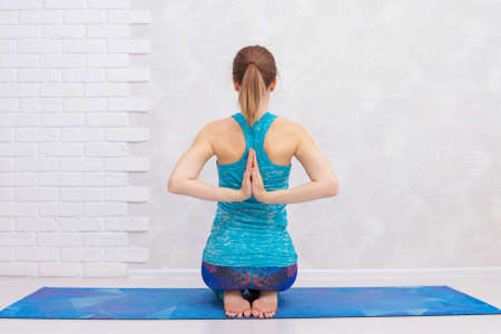 Portrait of beautiful young woman practicing yoga indoor. Woman in blue sportswear practice in light class. Horizontal, white background with brick wall. Copy space, back view.の写真素材