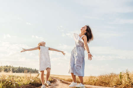 Beautiful young mother and her daughter having fun and jumping at the country road. Sky background. White clothes, freedom concept.の写真素材