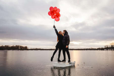 Love couple with red balloons kiss outdoor on sky and ice river background. Sunset light, black casual clothes.の写真素材