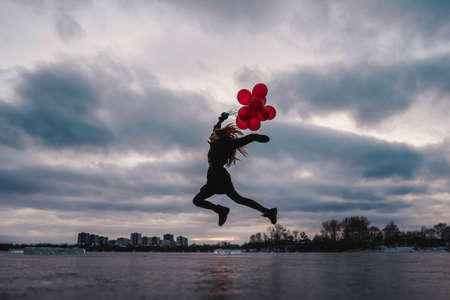 Imagination, happy girl jumping with red balloons at sunset on the frozen river, follow your dream. Copy space. Winter greeting card.の写真素材