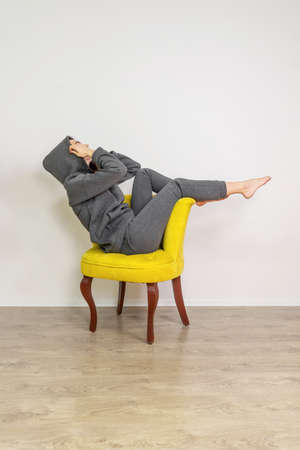 Young woman wearing gray costume on yellow chair on white background studio shot. Upside down. Minimal concept, gray and yellow colors 2021.の写真素材