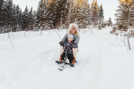In winter, happy mom and son ride a snow scooter from the mountain in white snow. Sunny forest on background.の写真素材