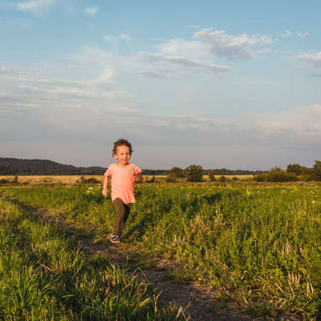 Little curly boy in orange t-shirt running in a meadow, sunset light. Beautiful landscape in summer, very light and happy scene.の写真素材