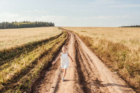 Little girl running on meadow in country road and throw little ears of corn . Sunset light, copy space.の写真素材
